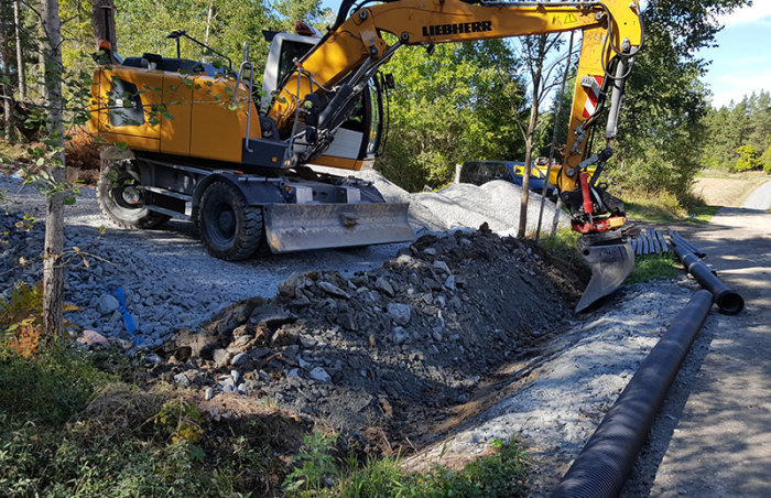 Grävmaskin i arbete med att gräva för vägtrummor, med grus och jordhögar runtomkring.