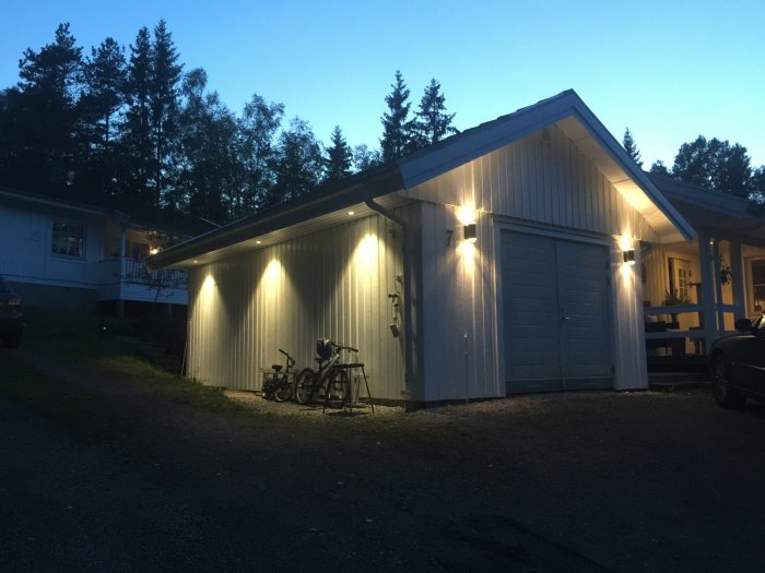 Vit garagebyggnad med ny utomhusbelysning i skymningen, cyklar parkerade vid sidan.