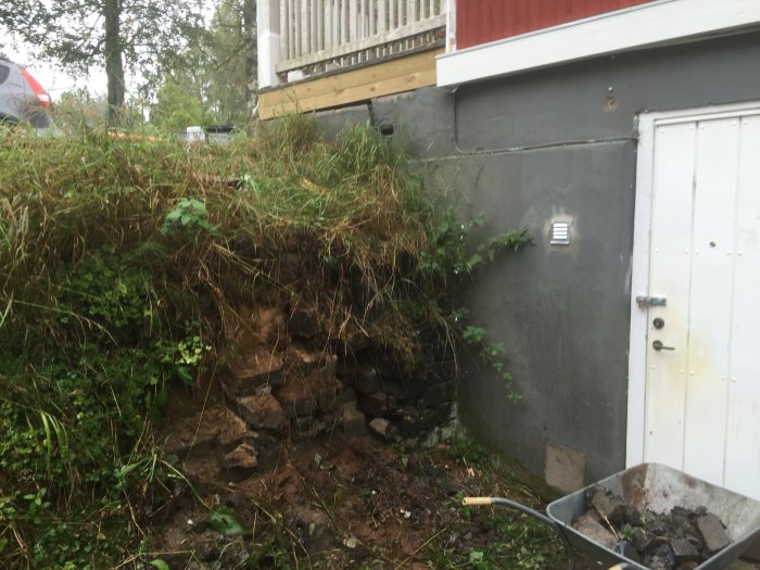 Skadad stödmur med lösa stenar och synlig erosion vid husgrund och veranda.