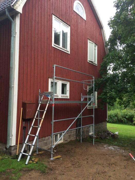 Estetisk grundmur av natursten synlig ovan jord vid ett rött hus med byggställning och ojämn gräsmatta.