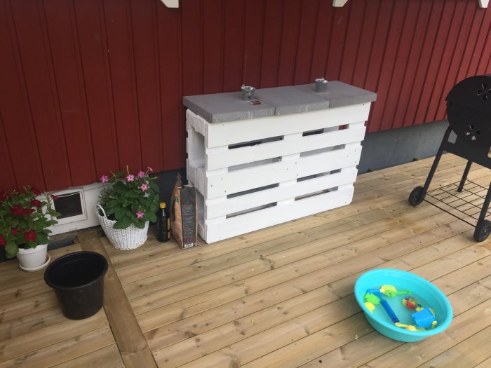 En nybyggd altan framför en röd husvägg med en vit uteköksbänk, blomkrukor, en grill och en barnpool.