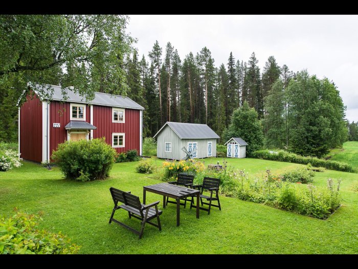 Rött hus med vita knutar och grön trädgård, följt av mindre byggnader och skog i bakgrunden.