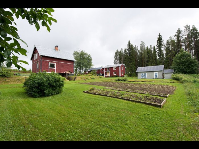 Ett rött hus med vita knutar och ett plåttak, omgivet av grönskande trädgård och byggnader på en lantlig tomt.