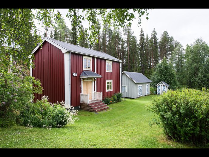 Rödmålat trähus med vita knutar och veranda, intill grönskande trädgård och två små uthus på en rymlig tomt.