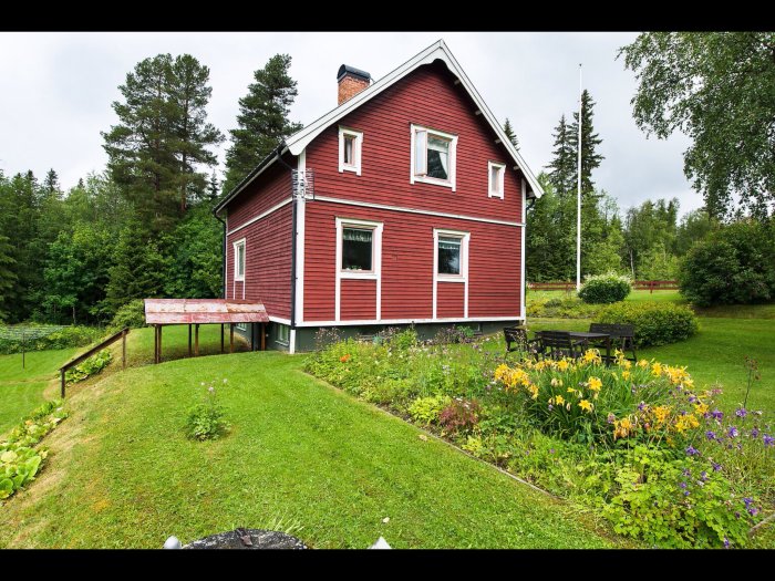 Tvåvånings rött hus med vita knutar, omgivet av en blommande trädgård och grönskande träd, under en molnig himmel.