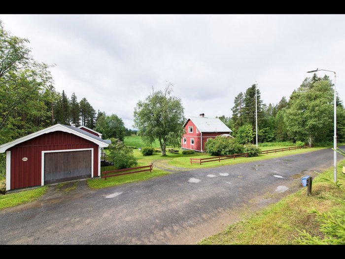 Röd tvåvåningshus med vita fönster, grått tak, grönskande omgivning, garage och staket.