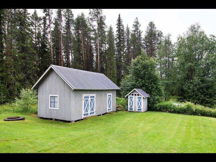 Ett gammalt grått trähus med blå dörrar och fönsterluckor, ett mindre skjul och naturtomt med träd i bakgrunden.