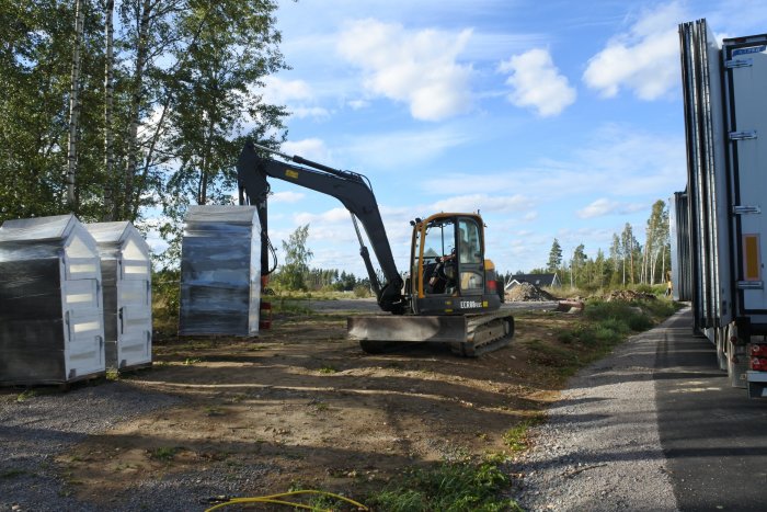 Grävmaskin i arbete på byggarbetsplats med avskalad mark och byggmaterial.