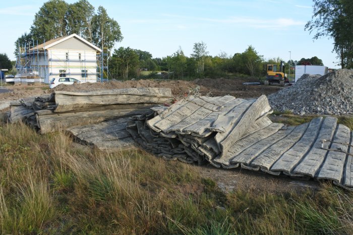 Byggarbetsplats med avtäckta markområden, hoprullade geotextilier och hus under uppbyggnad i bakgrunden.