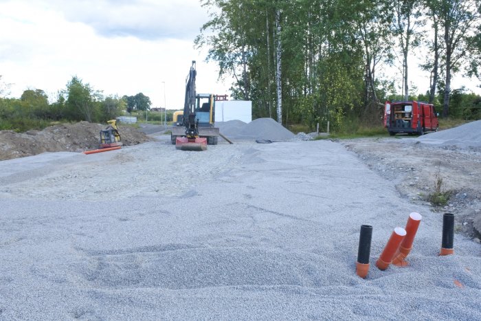 Byggarbetsplats med grävmaskin och högar av grus, jord samt koner på en avskalad tomt.