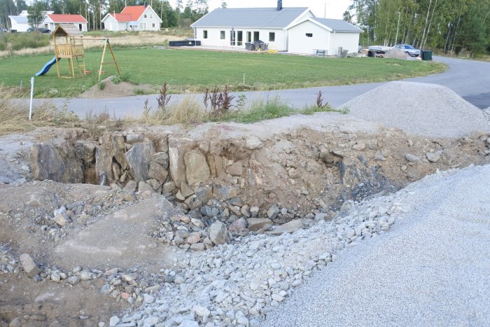 Nybyggd vit villa med grön gräsmatta i förgrunden och en uppgrävd tomt med grushög och synlig berggrund.