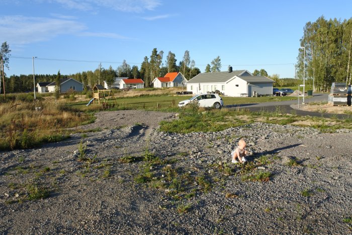 Tomt med grus och små växter, grävmaskinspår, nybyggda hus i bakgrunden, lekställning och parkerad bil.