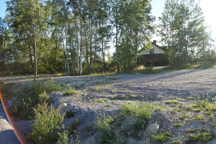 Avskalad tomt med grus och grässtrå, förberedd för byggprojekt, med träd i bakgrunden och ett hus.