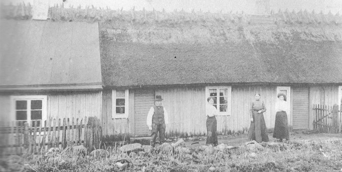 Svartvit bild från 1910-talet av träskomakarfamilj framför sitt hus med halmtak och träfasad.