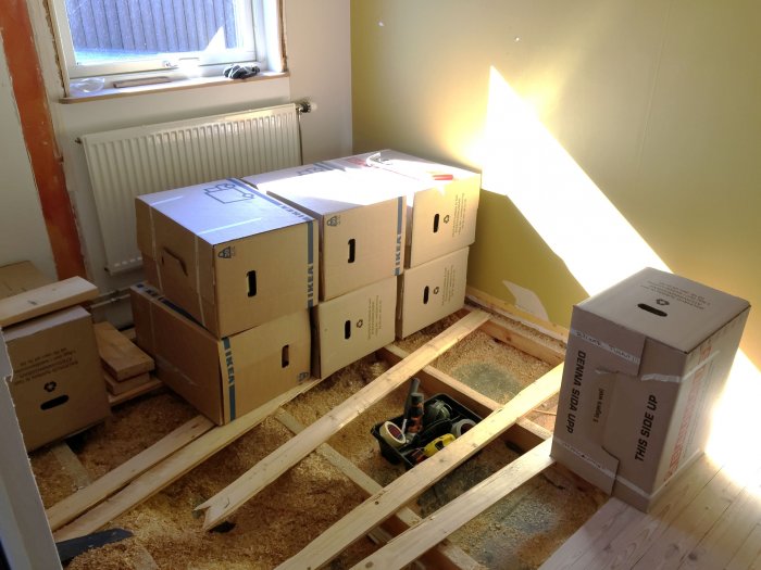 Flyttlådor arrangerade för att representera badkarets storlek i ett rum under renovering med träreglar och ospecificerad spånskiva på golvet.