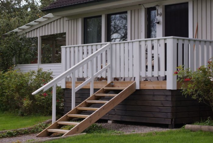 Nybyggd vit träaltan med trappa som ansluter till ett hus med ljusgrå fasad och mörkbrun grund.