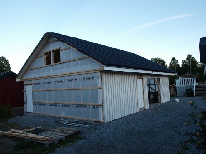 Nybyggd garagebyggnad med en sida grundad och målad, två dörrar monterade, och konstruktionsmaterial på marken.