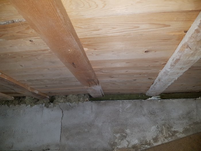 Träbjälkar i krypgrund med avvikande färger och skadad isolering längs betonggrund.