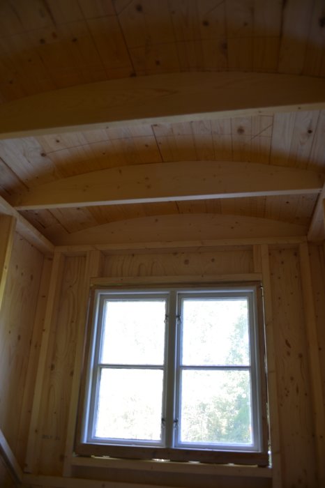 Interiörbild av råspontstak och väggar med synliga bjälklag ovanför ett fönster i ett pågående byggprojekt.