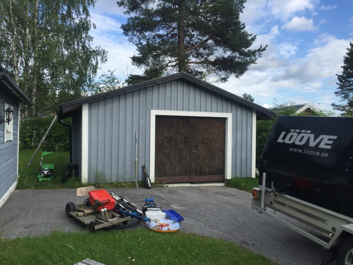 Före-bild av en garagebyggnad med gammal bruna dörrar och trädgårdsredskap och en släpvagn i förgrunden.