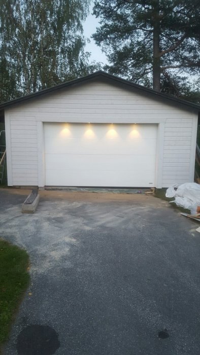 Bild på en vit garageport på en fristående byggnad med tre utvändiga lampor tända ovanför i skymningen.