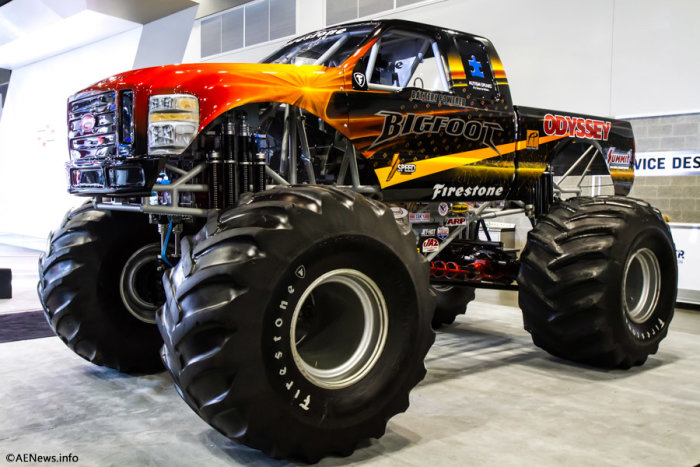 Orange-svart monstertruck med stora däck märkta 'Firestone' parkerad inomhus.