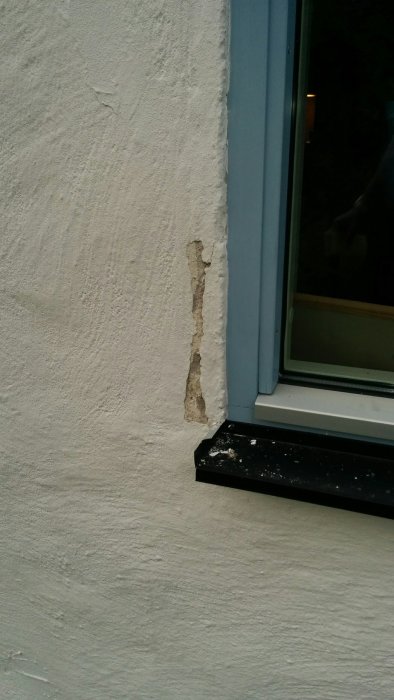 Skada på putsad fasad nära fönsterkarm där putsen har lossnat och exponerat underliggande material.