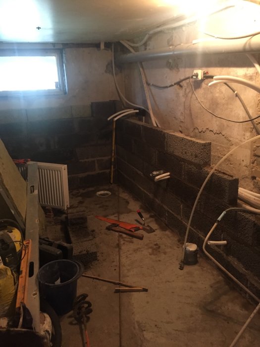 Påbörjad Leca-murning i källare med isolering och avloppsrör synliga.