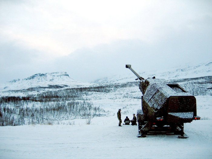 Ett gammalt artilleripjäs i ett snöigt landskap med tre personer som sitter vid pjäsens bas och samtalar.