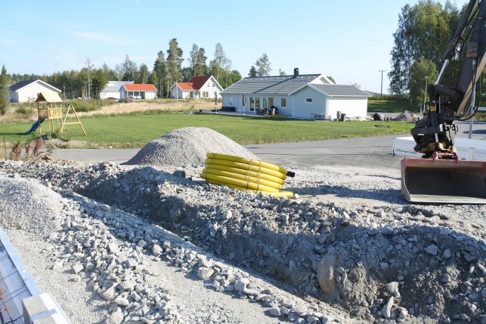 Tomt förberedd för gjutning med synliga avloppsrör i cellplast och grävmaskin i förgrunden, hus i bakgrunden.