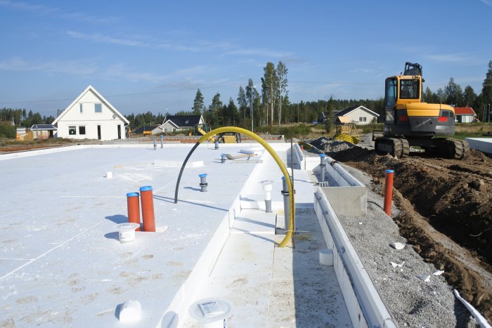 Tomt med förberedelser inför gjutning, synliga avloppsrör i cellplast, hus och grävmaskin i bakgrunden.