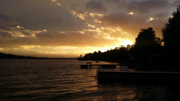 Solnedgång med dramatisk himmel över en brygga vid sjön omgiven av träd och båtar.