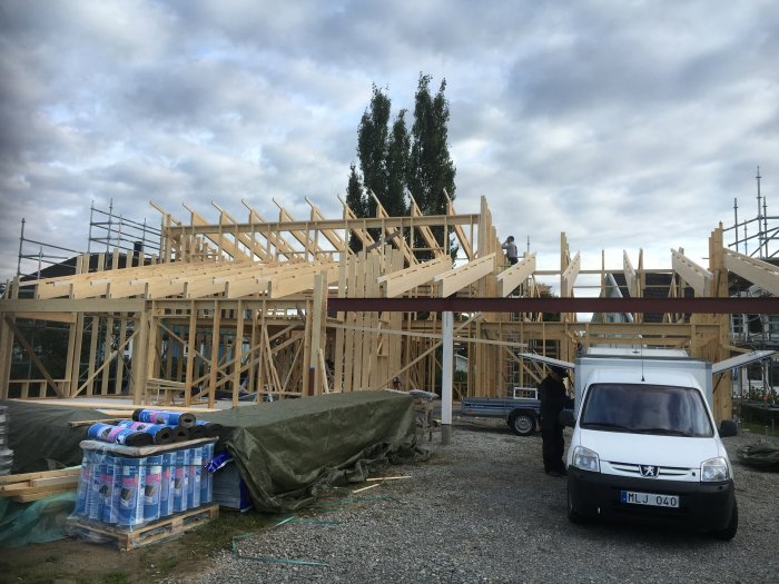 Takstolar på en carport och råspont på ett hus under konstruktion, byggmaterial och fordon syns framför.