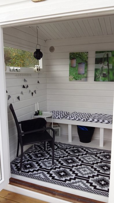 Inredd uteplats med svartvit matta, kuddar, bord och stol, vita väggar och svart hängande lykta.