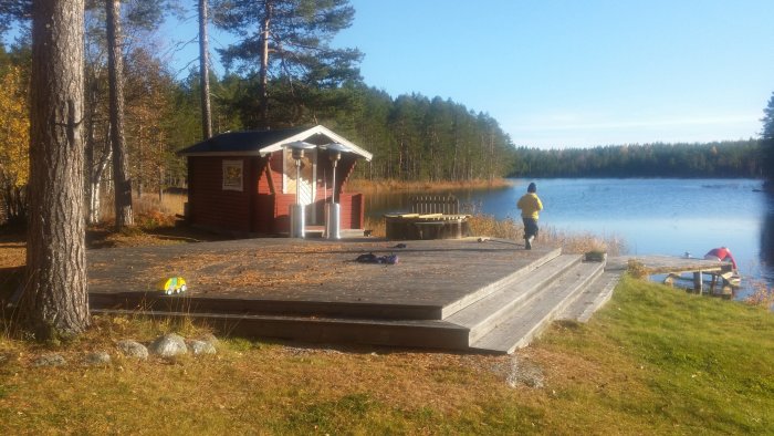 Solig utsikt över en stuga med altan nära en sjö, bastu och badbalja, person som går på bryggan.