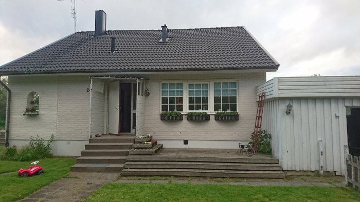 Ett vitmålat hus med svart tak och nyrenoverat fönsterparti med blomlådor, och en röd barnbil framför.
