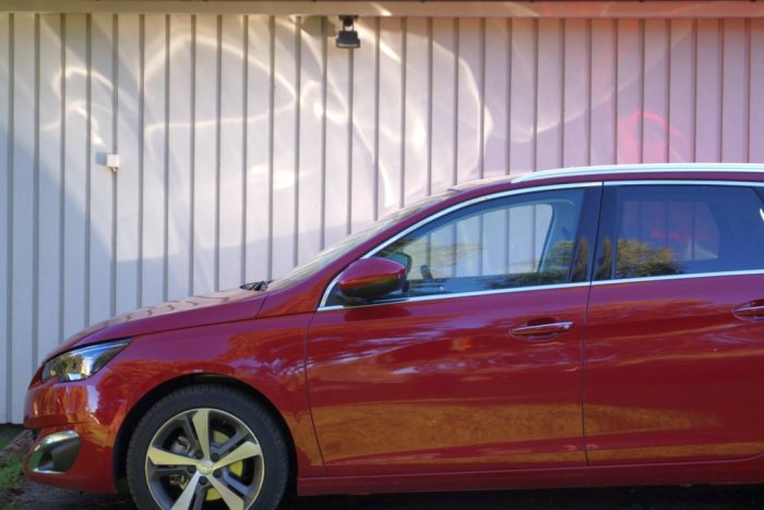 Röd bil parkerad framför ett hus med vågformade skuggor på fasaden.
