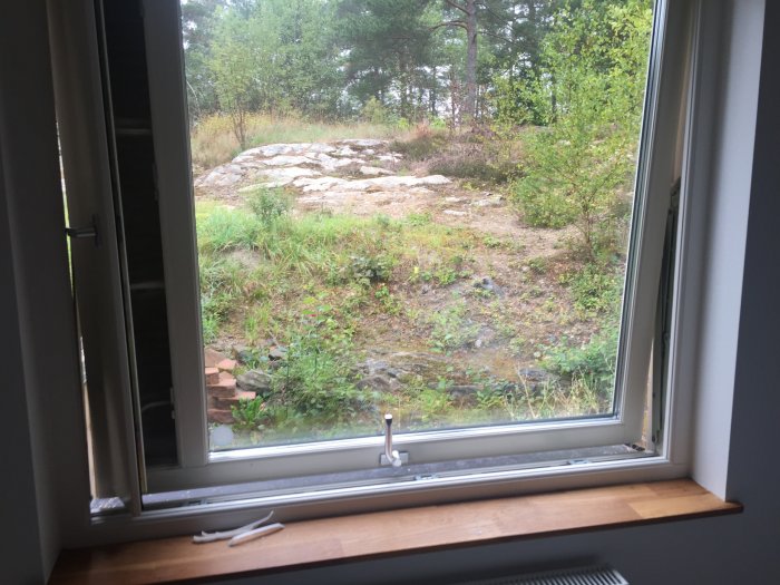 Fönster öppet inåt med ventilationslucka och utsikt över natur, spår av tidigare spärrar syns.