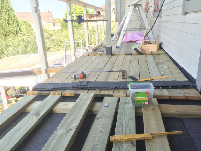Pågående arbete med att lägga trall på en balkong, med verktyg, träplank och delvis lagd trall synlig.