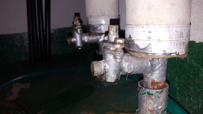 Äldre radiatorventiler och utloppsdetaljer med små droppar och korrosion, belägna nära en golvbrunn.