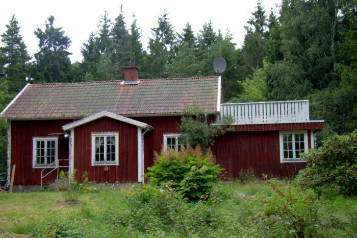 Rött trähus med slitet tegeltak som behöver renoveras, omgiven av träd och grönska.