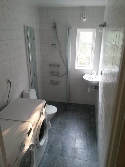 Renoverat badrum med dusch, handfat, WC och tvättmaskin, vita kakelväggar och mörkgrå klinkergolv.