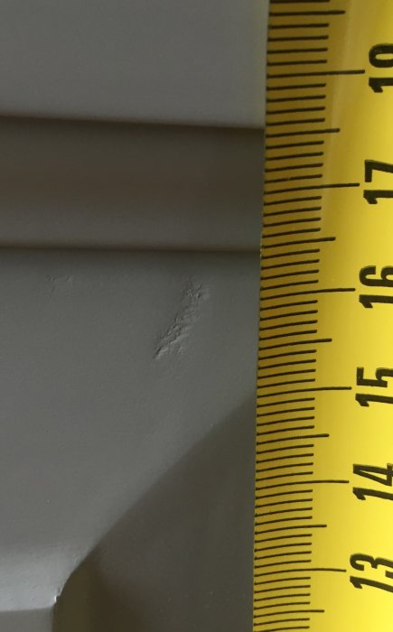 Ytlig defekt på gråmålad snickeriyta med mätsticka bredvid som visar storleken på skadan.
