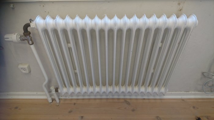 Vit sektionsradiator med termostat monterad på vägg, några tecken på rost vid basen.