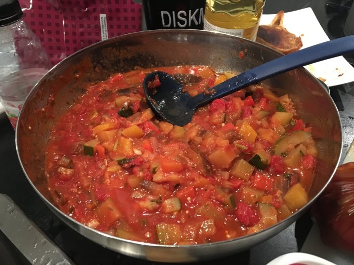 En hemlagad ratatouille i en stekpanna med en slev, tomater och squash syns tydligt.