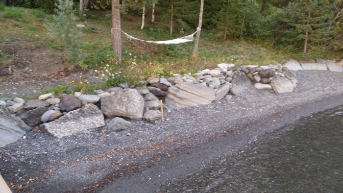 Strand med stora stenar och grus nära vattenkanten, hängmatta mellan träd i bakgrunden.