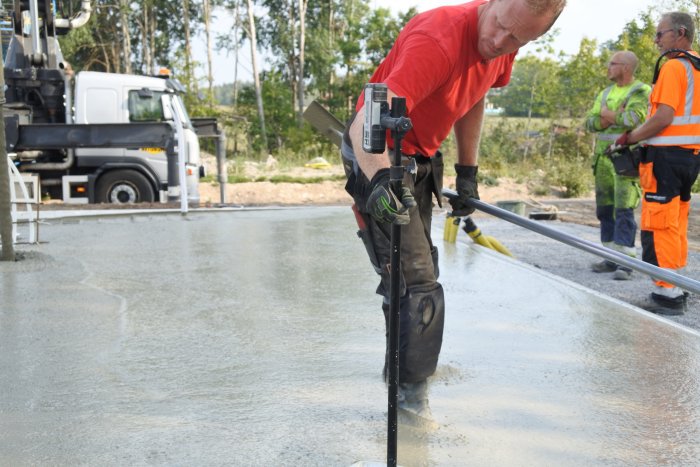 Arbetare i röd tröja som jämna till en nyligen gjuten betongyta medan kollegor observerar.