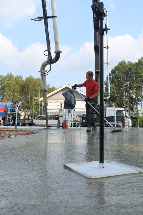 Arbetare i röd tröja jämnar ut nyligen gjuten betong med en spatel nära en betongpump på en byggarbetsplats.