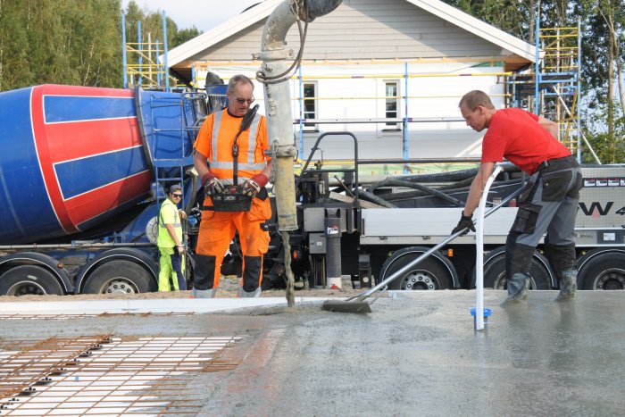 Arbetare syns gjuta en betongplatta med hjälp av betongbil och slang på en byggarbetsplats.