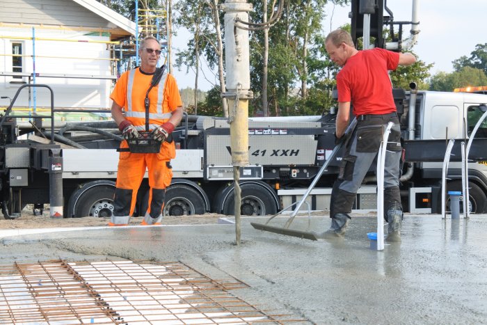 Två arbetare gjuter betong, en dirigerar en betongpump medan den andre jämnar ut betongen.